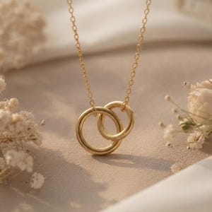 May include: A gold-toned necklace featuring a delicate chain and two interlocking circular rings as a pendant. The metal has a smooth, polished finish, and the design consists of one larger ring connected to a slightly smaller ring in a simple link.