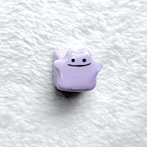 May include: A light purple, glossy, custom mechanical keyboard keycap shaped like a blob character with a simple, hand-painted black face featuring two small circular eyes and a thin, horizontal line for a mouth, set against a white textured surface.