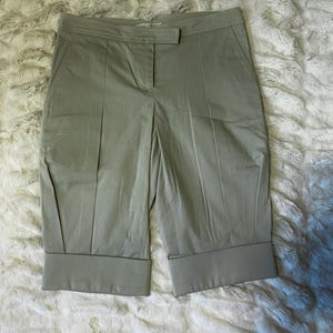 May include: Light olive green knee-length shorts featuring a structured waistband with a single button closure, a front zipper fly, and vertical seam detailing running down the front of each leg. The shorts have wide, folded cuffs at the hem.