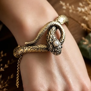 May include: A gold-toned metal bracelet shaped like a coiled snake with textured scales and a small white pearl set into the head. The snake body wraps around the wrist, featuring a slender metal chain hanging from the side of the bracelet.