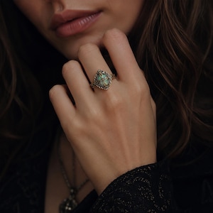 May include: A gold-toned ring featuring an oval-shaped, light green stone set within an intricate, filigree-style metal frame. The band displays a textured, braided pattern that extends from the central setting, creating a detailed appearance.