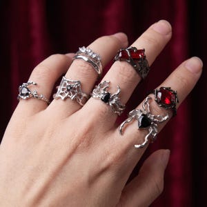 May include: A collection of six silver-toned rings worn on fingers, featuring intricate organic and web-like metalwork. The rings are set with various faceted gemstones, including deep red rectangular and oval stones, and black teardrop-shaped stones.