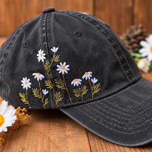 May include: A charcoal gray baseball cap featuring embroidered white daisy flowers with yellow centers and green stems. The embroidery is positioned on the side panel of the cap, showcasing detailed stitching of the floral stems and petals.