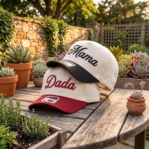 May include: Two baseball caps stacked on each other. The top cap is off-white with a black brim and the word Mama embroidered in black script. The bottom cap is off-white with a maroon brim and the word Dada embroidered in maroon script.