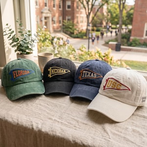 May include: Four baseball caps in green, black, navy blue, and off-white. Each cap features an embroidered pennant design with the text Florida, Michigan, Texas, or USC. The off-white cap also includes the text GAME DAY on the side panel.