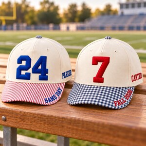 May include: Two off-white baseball caps with embroidered numbers. The left cap features a blue number 24, blue text reading PARKER and GAME DAY, and a red-and-white striped brim. The right cap has a red number 7, red text reading CARTER and FALL 2026, and a blue-and-white gingham brim.