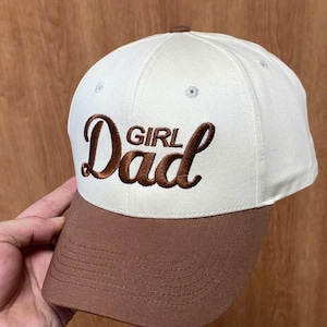 May include: A baseball cap with an off-white crown and a brown brim. The front of the cap features the embroidered text "Girl Dad" in a brown, cursive script font. The cap includes a button on the top center and visible stitching on the brim.