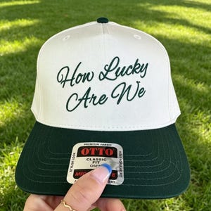 Op de afbeelding: Een witte honkbalpet met een donkergroene klep en de donkergroene geborduurde tekst How Lucky Are We op de voorkant. Een rechthoekige sticker met de merknaam OTTO en de woorden Premium Fabric Classic Fit is op de klep bevestigd.