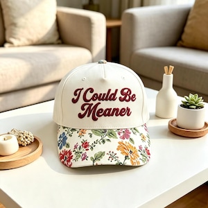 May include: A cream-colored baseball cap featuring the embroidered text "I Could Be Meaner" in dark red script on the front panel. The brim of the cap is decorated with a colorful floral pattern consisting of red, yellow, blue, and green flowers.