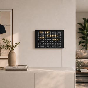 Könnte beinhalten: Eine rechteckige, schwarze Wortuhr an der Wand, die die Uhrzeit auf Deutsch über ein Gitter aus beleuchteten Buchstaben anzeigt. Das Zifferblatt besteht aus weißen Buchstaben auf schwarzem Grund, wobei einzelne Wörter gelb leuchten.