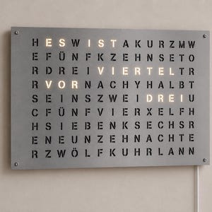 Könnte beinhalten: Eine rechteckige graue Wanduhr mit einem Gitter aus deutschen Buchstaben. Das Zifferblatt zeigt beleuchtete Buchstaben, die die Uhrzeit anzeigen, wobei die Wörter VOR, VIERTEL und DREI hervorgehoben sind, was Viertel vor drei bedeutet.
