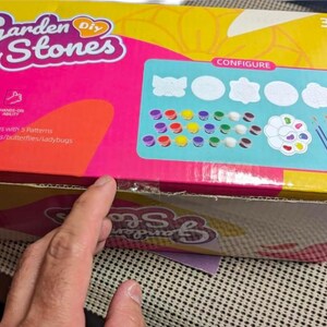 May include: A rectangular cardboard box for a Garden DIY Stones craft kit. The box is yellow and pink with the text Garden DIY Stones. It displays five white stone shapes, multiple small paint pots, a paint palette, and two paint brushes.