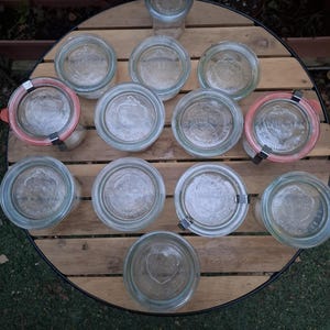 May include: A collection of twelve clear glass Weck jars arranged in a circular pattern. Each jar features a circular glass lid embossed with the Weck logo. Some jars include orange rubber gaskets and metal clips to secure the lids in place.