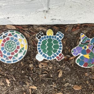 May include: Three decorative mosaic garden stones featuring a circular mandala pattern with red, blue, green, and yellow tiles, a turtle shape with a green shell and blue limbs, and a unicorn head silhouette with multicolored glass tile accents.