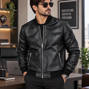 Peut inclure: Un blouson bomber noir avec un motif texturé en peau de serpent, une fermeture éclair argentée, deux poches latérales zippées, ainsi que des poignets et un ourlet côtelés noirs. Il est porté sur un t-shirt noir avec un pantalon noir et une montre.