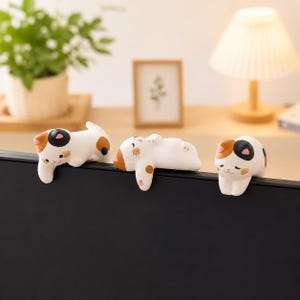 May include: Three small, white, calico cat figurines with orange and black patches are perched along the top edge of a black monitor. Each cat has a distinct pose, with painted facial features including closed eyes and pink accents on the ears.