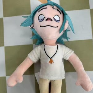 May include: A plush doll featuring a character with spiky teal hair, pale skin, and large white eyes. The doll wears a white short-sleeved shirt with a black necklace featuring an orange pendant, paired with light tan pants. The figure has a soft texture.