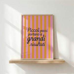 May include: A rectangular framed print featuring vertical stripes in alternating shades of pink and orange. Centered on the print is the black text "Piccoli passi portano a grandi risultati" written in a combination of serif and script fonts.