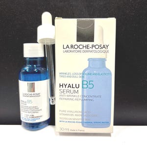 May include: A blue glass bottle of La Roche-Posay Hyalu B5 serum with a white dropper cap, accompanied by its white cardboard box. The packaging features the text: La Roche-Posay, Hyalu B5 Serum, Anti-Wrinkle Concentrate, Repairing Replumping, 30 ml.
