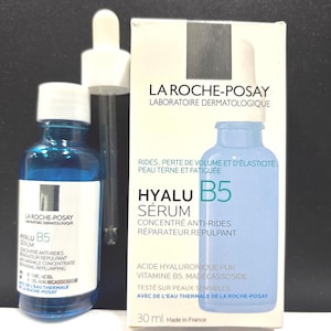 May include: A blue glass bottle of La Roche-Posay Hyalu B5 serum with a white dropper cap, accompanied by its white cardboard packaging box. The box features the product name, ingredients like hyaluronic acid and vitamin B5, and 30 ml volume.