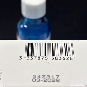 May include: A white rectangular box featuring a black barcode with the number 3 337875 583626 printed below it. Below the barcode, the text 54Z317 and 03-2028 are visible. A blue bottle with a white cap is positioned behind the box.