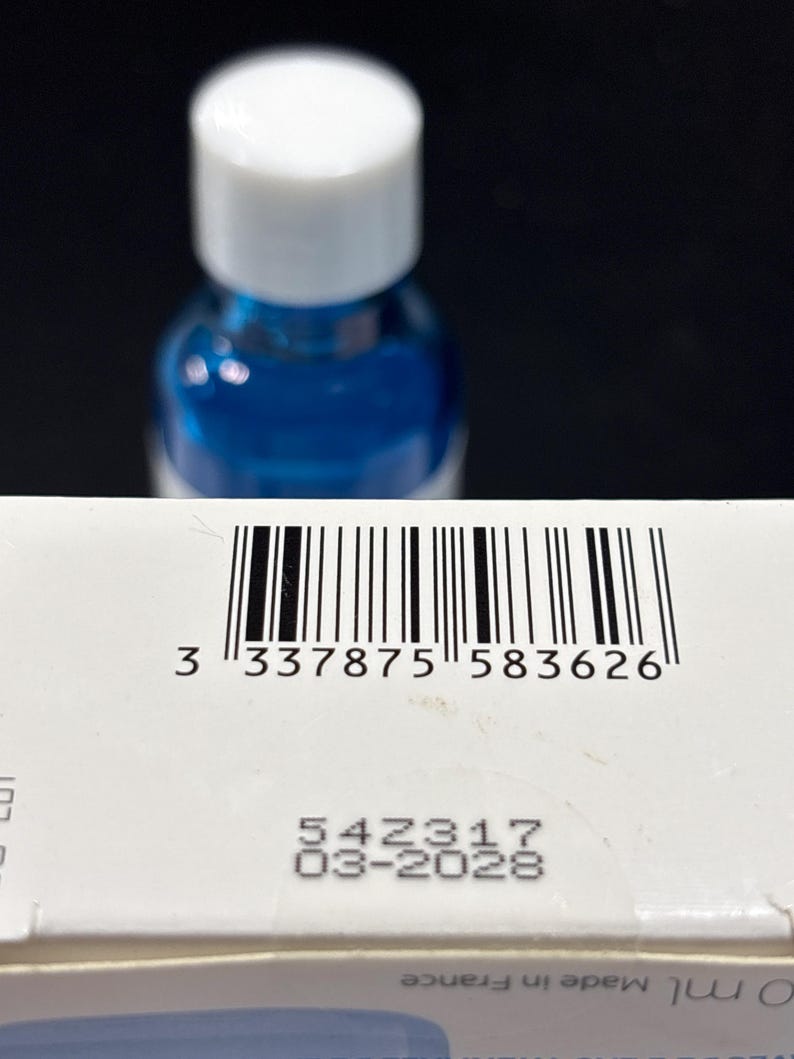 May include: A white rectangular box featuring a black barcode with the number 3 337875 583626 printed below it. Below the barcode, the text 54Z317 and 03-2028 are visible. A blue bottle with a white cap is positioned behind the box.