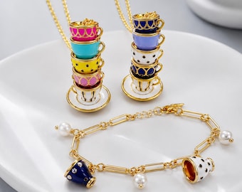 May include: Two gold-tone necklaces featuring pendants of stacked, colorful enamel teacups on saucers, alongside a gold-tone chain bracelet adorned with dangling charms of white pearl beads and miniature enamel teacups in blue and white polka dots.