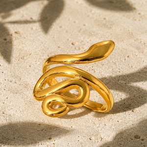 May include: A gold-toned ring shaped like a coiled snake. The metal band features a smooth, polished finish and curves into a continuous loop that mimics the form of a serpent with a rounded head and a tapered tail, creating a wrap-around design.