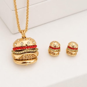 May include: A gold-toned necklace and matching stud earrings set featuring a hamburger pendant and charms. The burger design includes layers of clear crystals, red stones, and green stones, all set in a polished gold-colored metal finish.