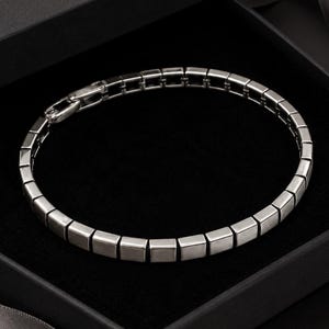 May include: A silver-toned bracelet composed of a series of connected square-shaped metal links. The bracelet features a polished finish and a secure clasp mechanism, arranged in a circular formation within a dark, square-shaped presentation box.