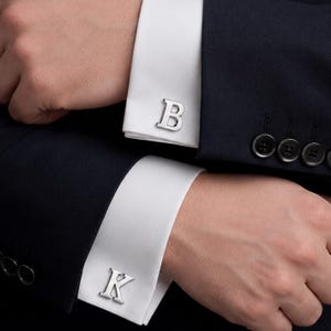 May include: A pair of silver-toned metal cufflinks featuring the letters B and K, attached to the white cuffs of a dark navy blue long-sleeved shirt. The cufflinks are polished and have a metallic finish, securing the folded fabric of the shirt sleeves.