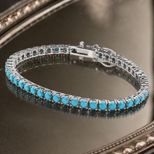 Op de afbeelding: Een zilverkleurige tennisarmband met een doorlopende rij ronde, helder turquoise stenen gezet in gepolijste metalen klauwen. De armband is voorzien van een veilige baksluiting met een zijdelingse veiligheidsklep voor een uniforme cirkelvorm.