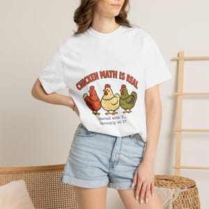 Op de afbeelding: Een wit T-shirt met korte mouwen en een afbeelding van drie kippen in rood, geel en groen. Boven de kippen staat de tekst "CHICKEN MATH IS REAL" in het rood en eronder staat de tekst "started with 3... currently at 17".