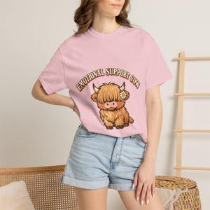 Op de afbeelding: Een lichtroze T-shirt met korte mouwen en een afbeelding van een pluizige bruine Schotse hooglander met een klein wit madeliefje op zijn kop. De tekst "EMOTIONAL SUPPORT COW" staat in een gebogen lettertype boven de illustratie.