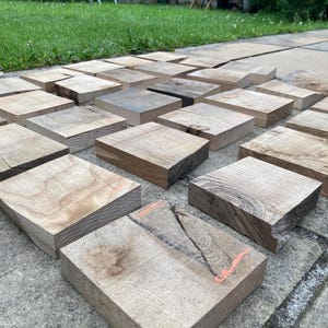 May include: A collection of square, unfinished wooden blocks arranged in a grid pattern. Each block displays natural wood grain, varying shades of light to dark brown, and unique textures. Some pieces feature visible knots, cracks, and faint orange markings.