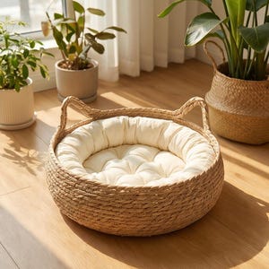 May include: A round pet bed constructed from woven natural seagrass fibers, featuring two upright ear-shaped accents on the rim. The bed includes a thick, tufted, cream-colored circular cushion that fits snugly inside the woven base structure.