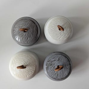Op de afbeelding: Vier ronde keramische potten met deksel, gerangschikt in een vierkant. Twee potten zijn donkergrijs en twee zijn wit. Elk deksel heeft een gestructureerd, stralend bladpatroon en een kleine, bruine, ringvormige handgreep op de bovenkant.
