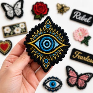 May include: A diamond-shaped embroidered patch featuring a central blue eye motif with gold and black detailing, surrounded by a scalloped border. The patch is displayed among various other embroidered patches including butterflies, flowers, and text.