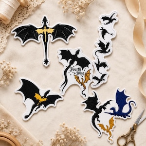 May include: A collection of embroidered patches featuring dragon silhouettes in black, gold, and blue. One patch displays the text Fourth Wing with small stars. The designs include various dragon shapes, some with wings spread, others in flight, and one vertical strip of small black dragons.