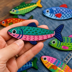 May include: A collection of embroidered fish patches in various colors including green, blue, red, and pink. Each patch features a detailed fish shape with stitched scales, fins, and eyes. The patches are arranged on a blue denim fabric background.