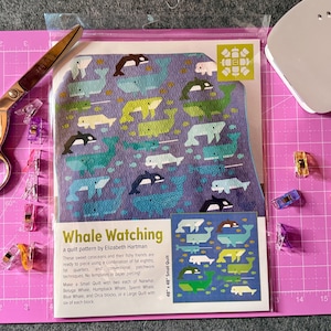 Könnte beinhalten: Eine Whale Watching Quilt-Anleitung von Elizabeth Hartman in einer Klarsichthülle. Das Cover zeigt einen violetten Hintergrund mit verschiedenen Wal-Illustrationen in Blau, Grün und Weiß. Der Text enthält den Titel und eine Größe von 122 cm.