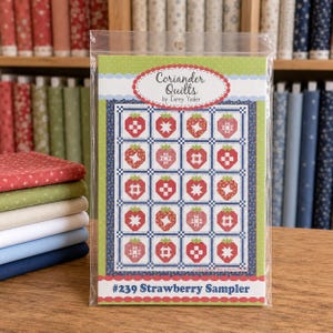 Può includere: Un libretto di modelli per trapunte intitolato #239 Strawberry Sampler di Coriander Quilts. La copertina presenta una griglia di venti blocchi a forma di fragola con motivi rossi e bianchi, su uno sfondo verde e un bordo blu floreale.