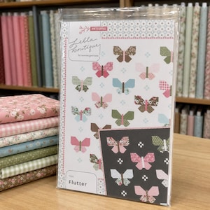 May include: A sewing pattern booklet titled Flutter by Lella Boutique featuring a white cover with a repeating butterfly motif in various colors and patterns, including a dark gray inset panel with pink and white butterflies, all inside a clear plastic sleeve.