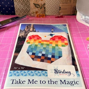 Könnte beinhalten: Ein Quilt-Anleitungsheft mit dem Titel Take Me to the Magic von Sterling Quilt Co. Das Cover zeigt eine regenbogenfarbene, pixelige Mausohren-Form auf weißem Hintergrund, ein Schlossbild und den aufgedruckten Text 76" x 76".