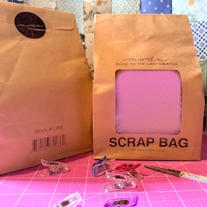 May include: Two brown paper bags labeled Moda, one with a window showing pink fabric. The bags are marked Scrap Bag, Net Weight Apx. 1/2 lb, and Stock # LBM. Several clear plastic sewing clips and a pair of floral scissors are scattered in front.