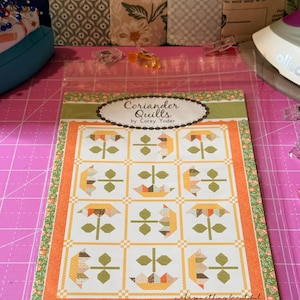 May include: A quilt pattern booklet titled Coriander Quilts by Corey Yoder #221 Sweetest. The cover features a grid of twelve floral blocks in orange, yellow, and green, surrounded by a green floral border and an orange frame on a white background.