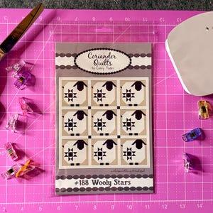 May include: A Coriander Quilts #188 Wooly Stars pattern booklet in a clear plastic sleeve, surrounded by metal scissors, a white iron, and several colorful plastic sewing clips on a pink cutting mat with a visible imperial grid.