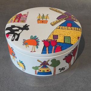 Pode incluir: Um recipiente de armazenamento redondo de cerâmica branca com ilustrações coloridas desenhadas à mão de moinhos de vento, uma vaca preta e branca, tulipas vermelhas e pequenas casas que decoram a tampa e as laterais do objeto.