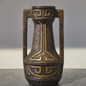 May include: A bronze-toned metal vase featuring a textured, hammered surface and two vertical side handles. The body is decorated with raised, smooth geometric patterns, including Greek key motifs around the neck and base, and a central vertical line.