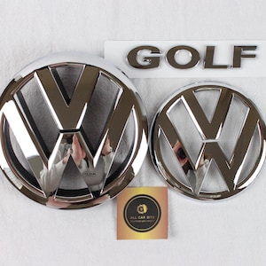 Könnte beinhalten: Zwei runde, verchromte Volkswagen-Embleme in verschiedenen Größen, ein verchromter GOLF-Schriftzug und eine kleine quadratische Karte mit dem Text ALL CAR BITZ. Die Embleme zeigen das VW-Logo mit einer polierten, reflektierenden Oberfläche.